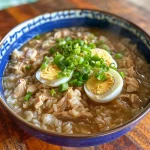 ['Close-up of rice porridge with garnishes and broth in a bowl.', 'A side view of creamy rice porridge topped with green onions and boiled eggs.', 'Juicy rice porridge in a bowl, showcasing its ingredients and texture.', 'Delicious Reisbrei served in a bowl, featuring onions and herbs.']