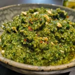 ['Side view of vibrant green Rosenkohl pesto in a bowl, showcasing its chunky texture.', 'Close-up image of freshly made Rosenkohl pesto with visible ingredients like basil and almonds.', 'Juicy and rich green Rosenkohl pesto spread thickly in a small dish, highlighting its freshness.', 'Detailed view of a creamy Rosenkohl pesto, garnished with basil leaves and almonds.']