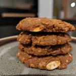 ['Close-up view of a Süßkartoffel cookie with a golden exterior and chocolate nibs.', 'Side view of a freshly baked Süßkartoffel cookie showcasing its chewy texture.', 'Juicy Süßkartoffel cookie featuring chunks of nuts and a sprinkle of cocoa nibs.', 'Delicious Süßkartoffel cookie on a plate, highlighting its rustic appearance.']