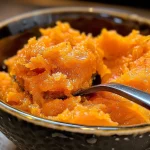 ['Close-up of a jar of sweet potato carrot jam, showcasing its vibrant orange color.', 'Side view of sweet potato carrot jam in a glass jar, highlighting the texture and richness.', 'A spoonful of sweet potato carrot jam with the jar in the background, emphasizing its glossy appearance.', 'Sweet potato carrot jam spread on a slice of bread, displaying its thick consistency.']