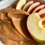 ['Close-up view of a creamy apple dip in a small bowl, with fresh apple slices beside it.', 'Side view of a creamy peanut butter and maple syrup dip served with sliced apples.', 'Juicy apple slices next to a rich, creamy dip made from peanut butter and maple syrup.', 'Appetizing apple dip in a bowl, showcased with vibrant apple slices for dipping.']