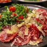 ['Close-up of thinly sliced figs arranged with prosciutto and arugula, garnished with pine nuts and shaved parmesan.', 'Side view of a vibrant plate featuring fig carpaccio, showcasing layers of ingredients including prosciutto and greens.', 'Detailed shot of fig carpaccio with drizzled olive oil, highlighting the colors and textures of figs and toppings.', 'Artistic presentation of Feigencarpaccio with fresh arugula, nuts, and cheese, captured against a neutral background.']