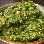 ['Close-up view of vibrant green Grünkohl pesto in a bowl.', 'Juicy Grünkohl pesto displayed elegantly with fresh herbs.', 'Side view of thick Grünkohl pesto surrounded by ingredients.', 'Freshly made Grünkohl pesto with a glossy texture in a rustic setting.']